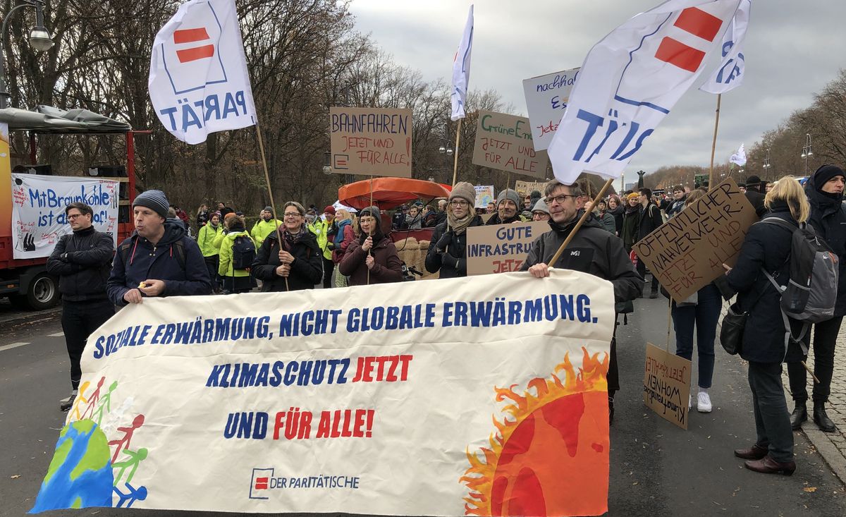 Foto mit Menschen auf einer Demonstration, die Schilder und ein Banner mit der Aufschrift "Soziale Erwärmung, nicht globale Erwärmung. Klimaschutz jetzt und für alle" halten.
