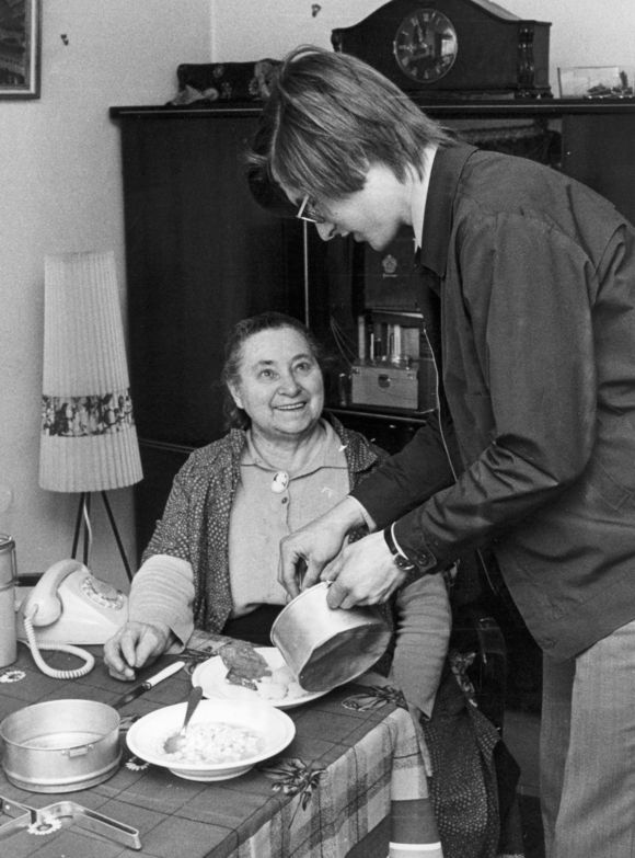 Ältere Fotoaufnahme: Eine ältere Frau lächelt einen jungen Mann an, der ihr Essen auf einen Teller aufträgt, die Frau sitzt in einem Wohnzimmer mit Telefon, Stehlampe und Schrankwand. 