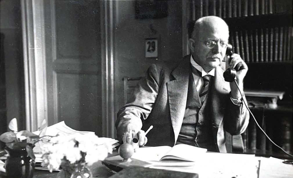 Sehr alt wirkendes Foto: Ein Mann sitzt telefonierend und mit einem Stift in der Hand an einem Schreibtisch vor einer Bücherwand. Der Schreibtisch ist voll mit Papieren. Der Mann hat eine Glatze, trägt eine kleine Brille sowie einen Schnurrbart sowie Hemd, Weste, Jackett und Krawatte.