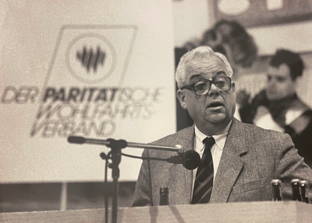 Foto von Dieter Sengling an einem Mikrofon stehend, im Hintergrund ist das frühere Logo des Paritätischen Gesamtverbandes zu sehen.