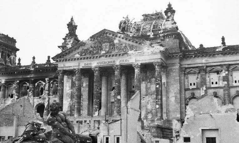 Historisches Foto des Berliner Reichstags, vor dem eine Trümmerlandschaft zu sehen ist, der Reichstag selbst sieht zerschossen und ausgebrannt aus.