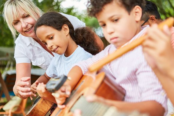 Kinder unterschiedlicher Haut- und Haarfarbe sitzen nebeneinander und üben, auf Gitarren zu spielen, eine Lehrerin schaut zwischen ihnen hervor und gibt Hilfestellungen.