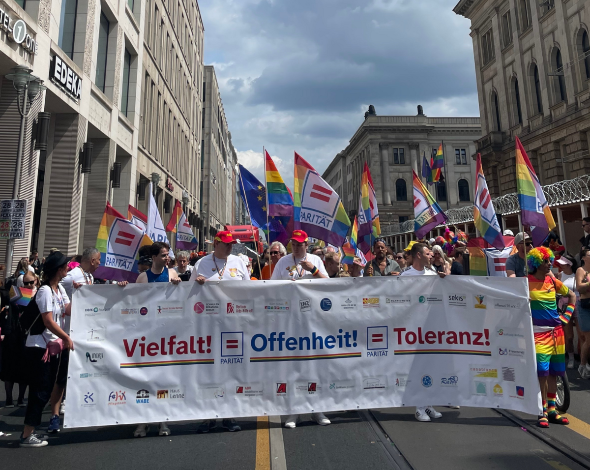 Foto mit vielen unterschiedlichen bunt gekleideten Menschen, die ein Banner in Regenbogenfarben halten mit der Aufschrift: "Vielfalt! Offenheit! Toleranz!".