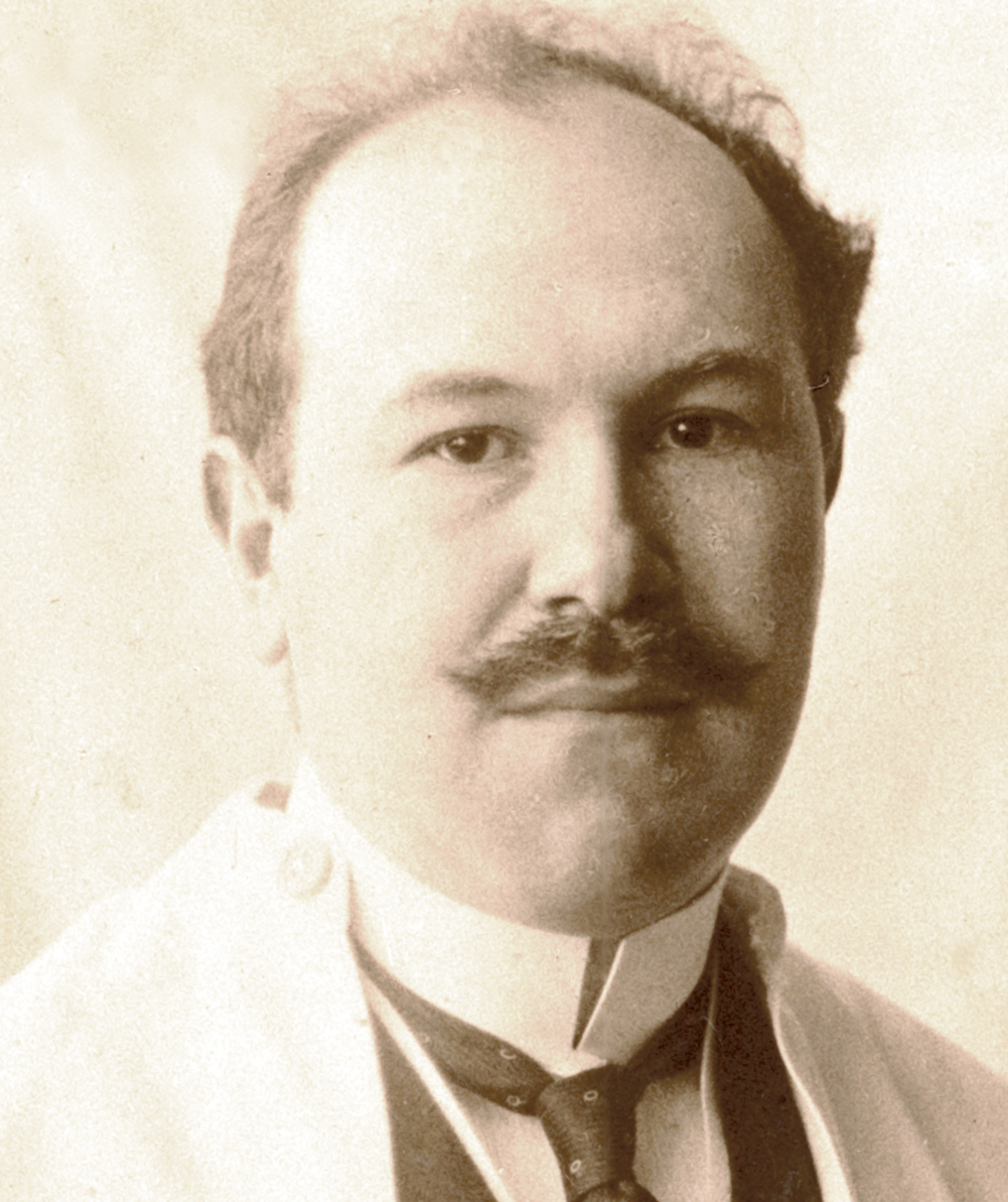 Ein sehr altes Foto eines Mannes mit Schnurrbart, Halbglatze und weißer Jacke in Hemd und Krawatte.