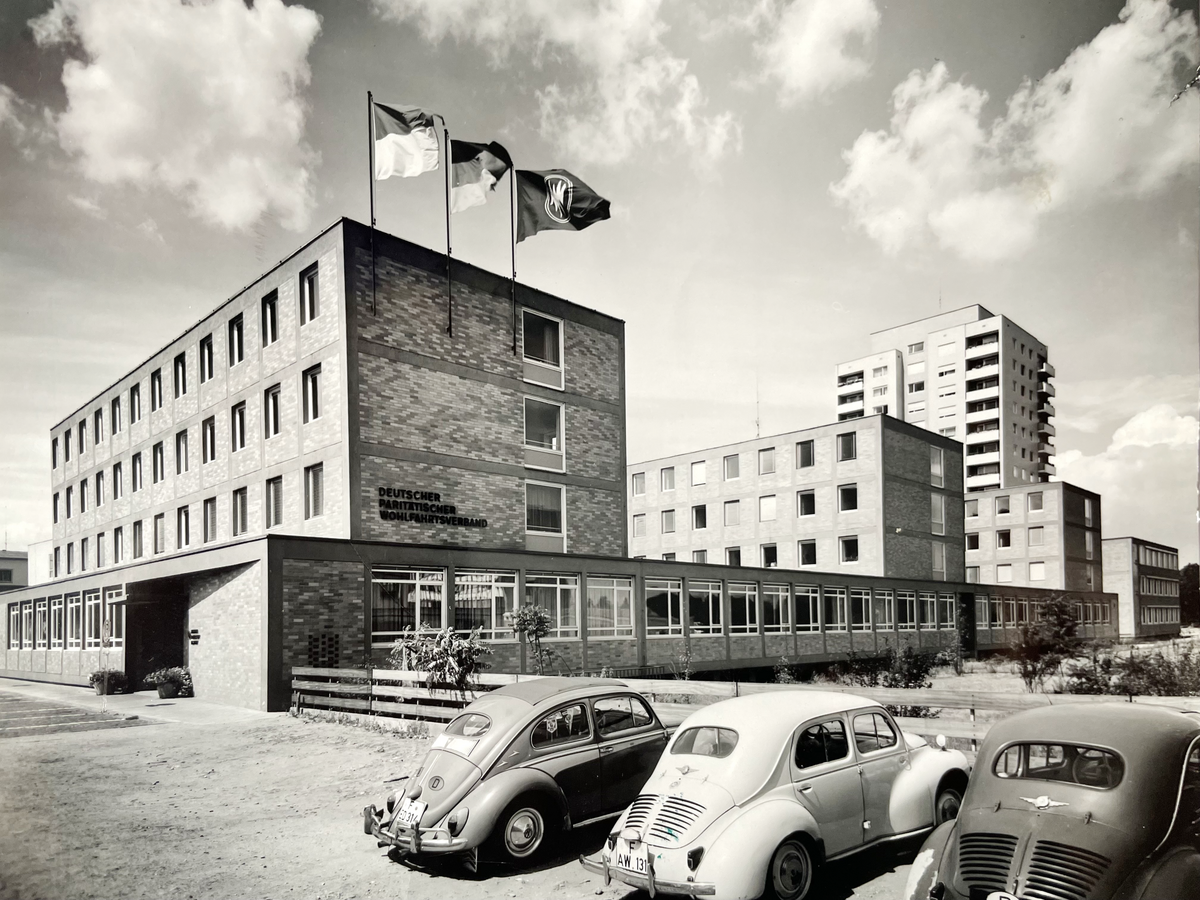Älteres Foto eines modernen Gebäudekomplexes bestehend aus drei Gebäuden, verbunden durch einen Flachbau, davor stehen drei VW-Käfer. Auf dem Gebäude wehen u.a. die Fahnen Deutschlands sowie des Paritätischen.