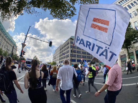 Auf einer Demonstration trägt ein Mann bei Sommerwetter eine größe Fahne mit dem Ist-Gleich-Zeichen des Logos des Paritätischen Wohlfahrtsverbandes, auf der Fahne steht: Parität.