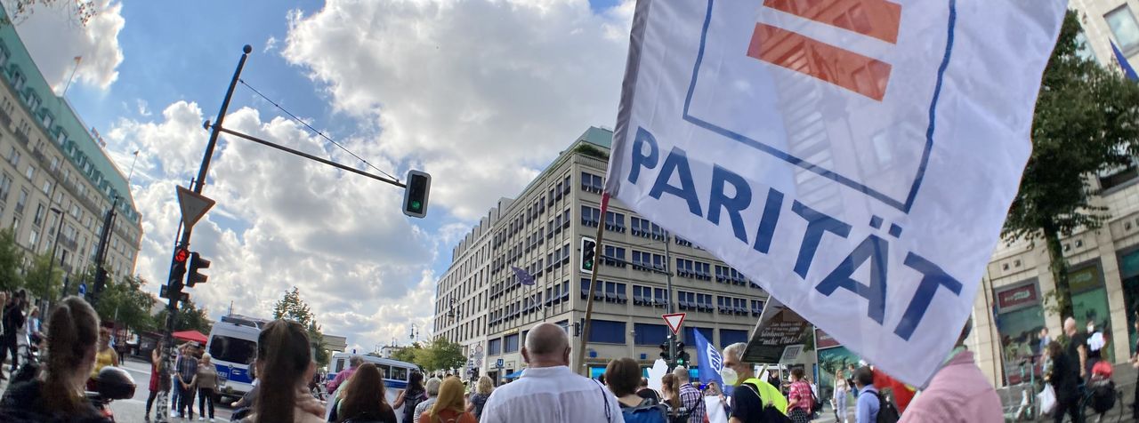 Auf einer Demonstration trägt ein Mann bei Sommerwetter eine größe Fahne mit dem Ist-Gleich-Zeichen des Logos des Paritätischen Wohlfahrtsverbandes, auf der Fahne steht: Parität.