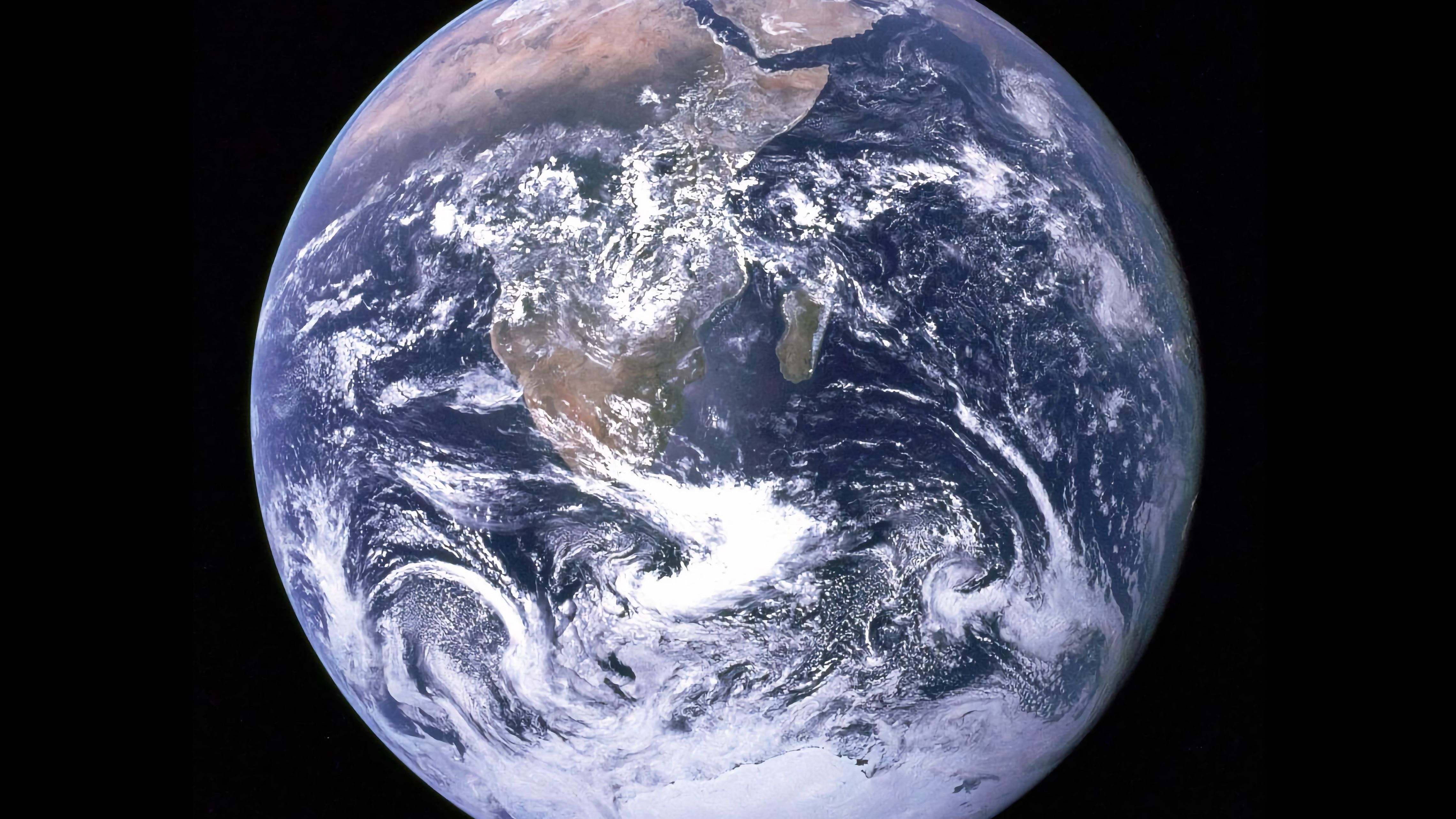 Aufnahme aus dem Weltall von der leuchtenden Erde auf der Meere, Wolken und Landmassen zu sehen sind, rund um die Erde ist der Hintergrund dunkel.  