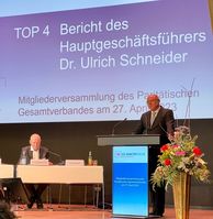 Ein Mann steht auf einer Bühne und spricht. Dahinter wird Text eingeblendet: Bericht des Hauptgeschäftsführers Dr. Ulrich Schneider.