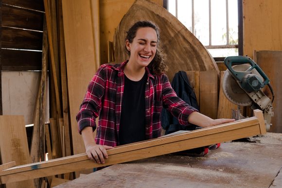 Eine Frau steht lachend mit einem Stück Holz vor einer Kreissäge. 