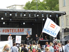 Auf einer Bühne vor einer Demonstration ist eine Banner "Unteilbar" zu sehen. Davor eine weiße Fahne mit dem Logo "Parität". 