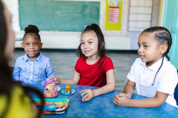 Drei Kinder sitzen in einem Kindergarten an einem Tisch und hören aufmerksam ihrer Erzieherin zu.