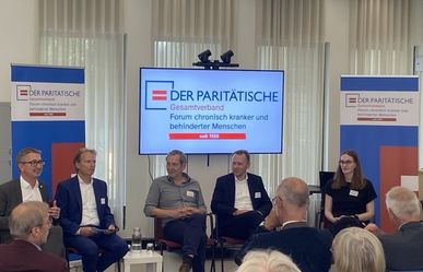 Eine weitere Podiumsrunde mit vier sitzenden Diskussionsteilnehmenden, gefilmt über die Schulter einer im Vordergrund sitzenden Person aus dem Publikum.
