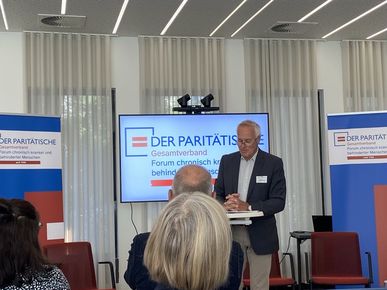 Ein älterer Mann in einem dunklen Sakko steht an einem Stehpult im Tagungsraum und spricht zu den Gästen. Im Hintergrund zeigt ein großer Bildschirm das Logo „DER PARITÄTISCHE Gesamtverband – Forum chronisch kranker und behinderter Menschen“.