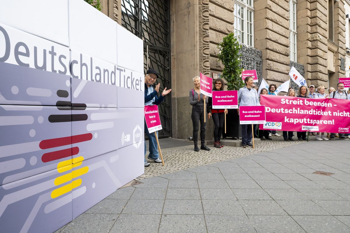 Zu sehen ist ein übergroßes Deutschlandticket aus Karton, das vor einem offiziellen Gebäude aufgestellt wurde. Daneben steht eine Gruppe von Menschen, die Banner und Schilder hochhalten, auf denen Protestbotschaften für den Erhalt des Deutschlandtickets zu lesen sind. Die Schilder sind pink mit weißer Schrift.