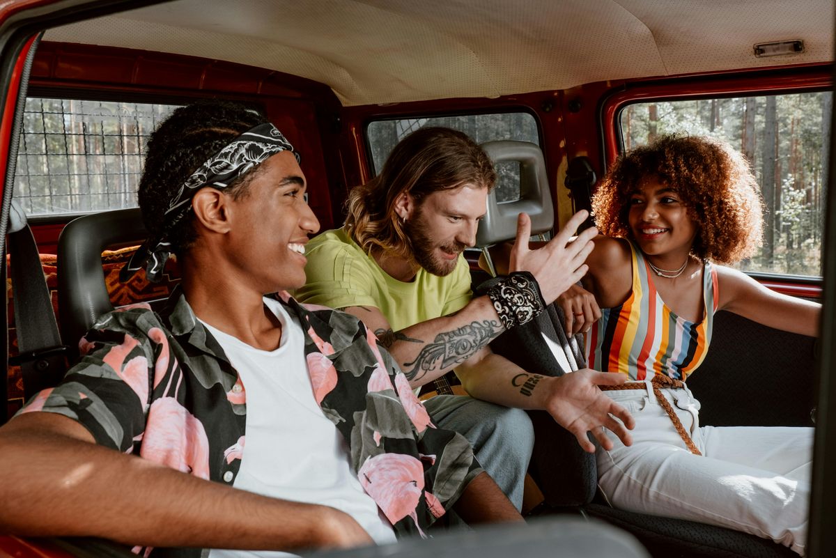 Drei junge Menschen, zwei Männer und eine Frau, mit bunten Outfits sitzen lachend in einem alten Auto.