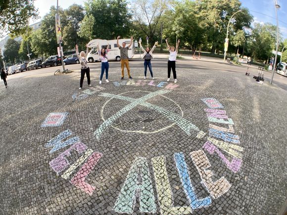 Eine Gruppe steht jubelnd vor einem Kreidebild: Auf die Straße ist ein großes Wahlkreuz gemalt plus der Text "Geh wählen, weil alle zählen!" 