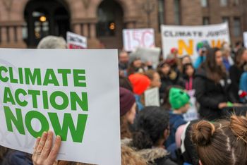 Das Bild zeigt ein Demoplakat mit der Aufschrift Climate Action Now. Im Hintergrund ist eine Gruppe demonstrierender Menschen zu sehen.