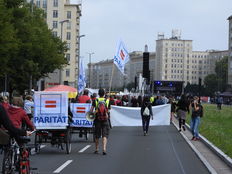 Viele Menschen auf einer Demonstration, die von hinten fotografiert wurde: Ein Rikscha-Fahrrad ist in einer weiße Fahne mit dem Logo "Parität" eingewickelt.