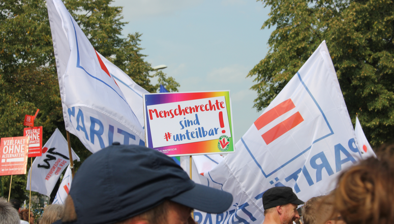 Foto einer Demonstration mit Fahnen, auf denen das Logo des Paritätischen zu sehen ist und einem Demo-Schild mit der Aufschrift: "Menschenrechte sind #unteilbar!".