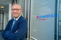 Ein Mann, mit Brille steht lächelnd vor einer Fensterscheibe, auf welcher das Logo des Paritätischen zu sehen ist. Es ist Prof. Dr. Rosenbrock, Vorsitzender des Paritätischen Gesamtverbandes.