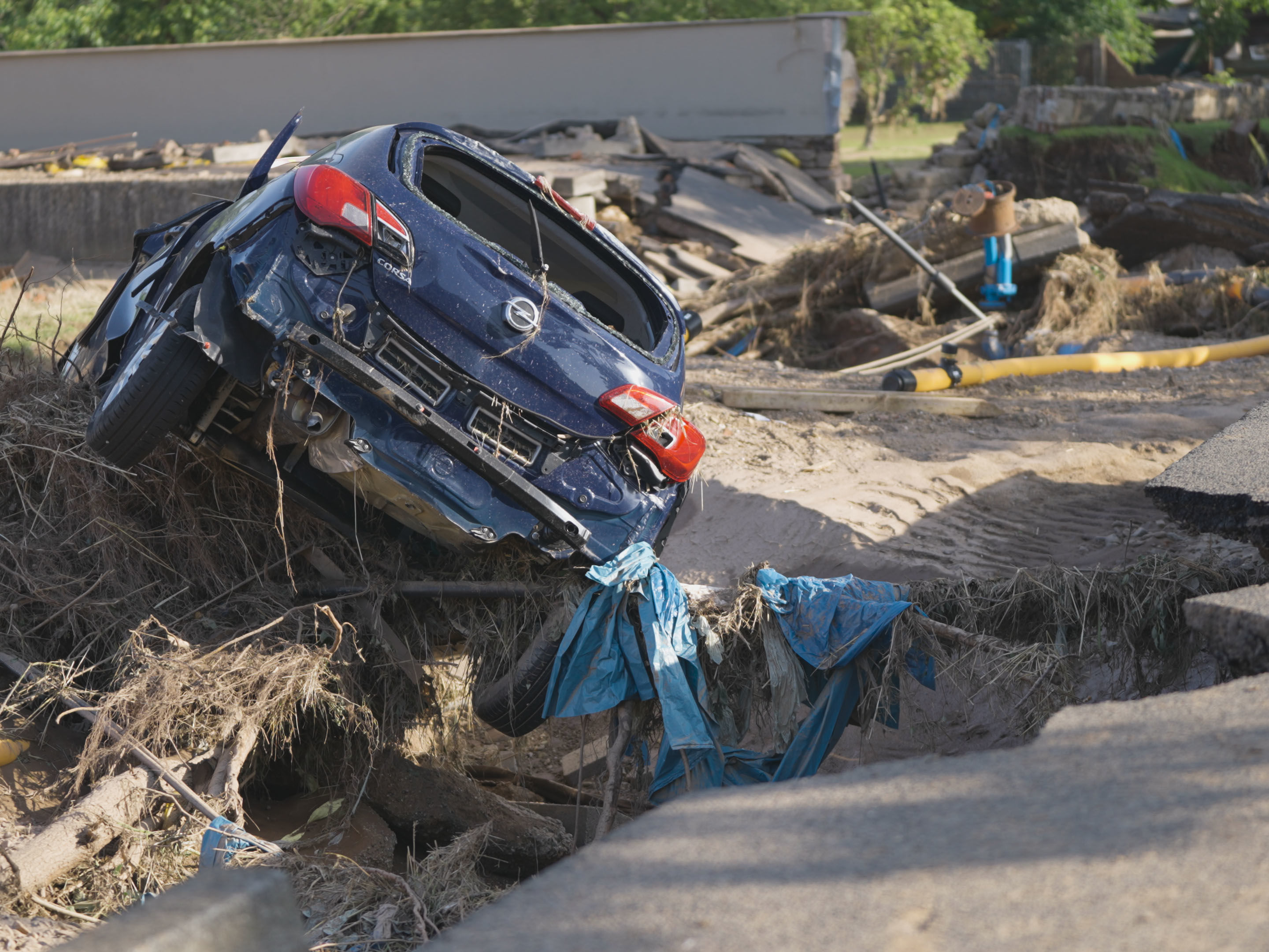 Eine Straße ist von der Flut aufgerissen, darauf steht ein zetrümmertes Auto, zu sehen ist außerdem Geröll, auf dem Kleidung liegt