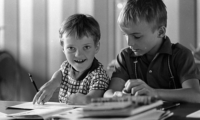 Altes Foto von zwei Kindern, die an einem Tisch an Schulheften sitzen. Ein Kind hat eine sichtbare körperliche Behinderung am Arm, das andere nicht.