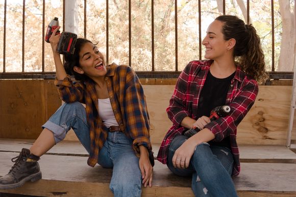 Zwei junge Frauen in Handwerkskleidung sitzen lachend auf einer Werkbank.