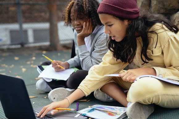 Zwei jugendliche Mädchen sitzen mit Stiften und Papier vor einem Laptop und erledigen Hausaufgaben. 