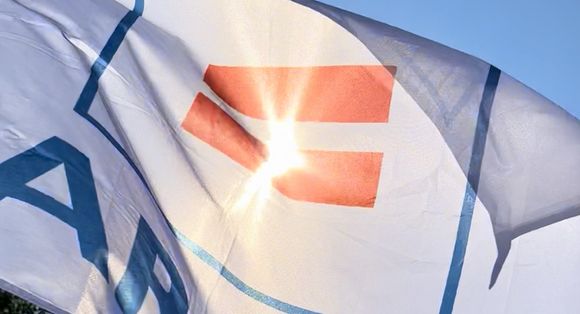 Eine Fahne mit dem Logo des Paritätischen, einem Gleichheitzszeichen, weht im Wind, dahinter scheint die Sonne durch das Logo.