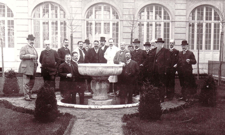 Auf einem sehr alten Foto steht eine Gruppe von Männern in offizleller Kleidung um einen Brunnen, viele haben Schnurrbärte und tragen Hut.