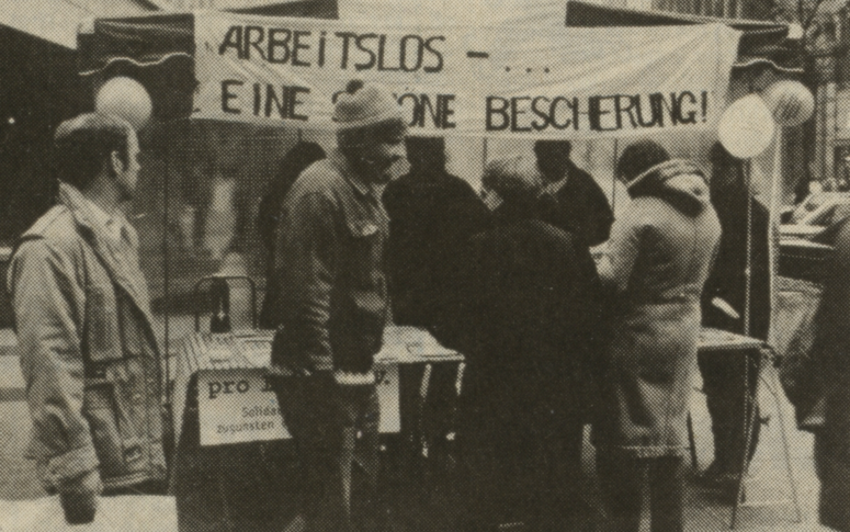 Foto auf dem sich Menschen vor einem Stand versammeln über dem ein banner mit der Aufschrift "Arbeitslos - Eine schöne bescherung" steht.