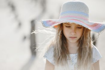 Ein etwa sienjähriges Mädchen mit langen blonden Haaren und einem große Hut schaut traurig auf den Boden. 