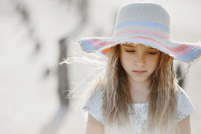Ein etwa sienjähriges Mädchen mit langen blonden Haaren und einem große Hut schaut traurig auf den Boden. 