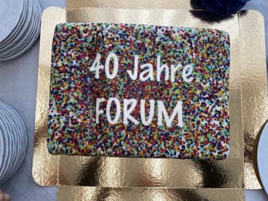 raufsicht auf einen rechteckigen Jubiläumskuchen mit buntem Streuselbelag auf einer goldenen Unterlage und dem weißen Schriftzug „40 Jahre FORUM“.