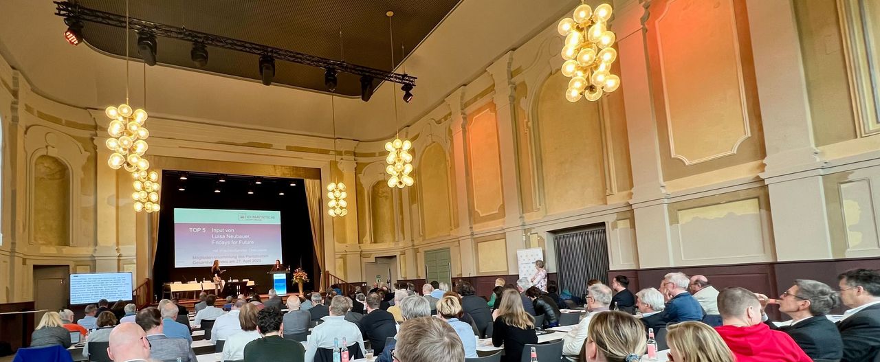 Viele Menschen sitzen gemeinsam in einem festlichen Saal, auf der Bühne sprechen zwei Frauen.