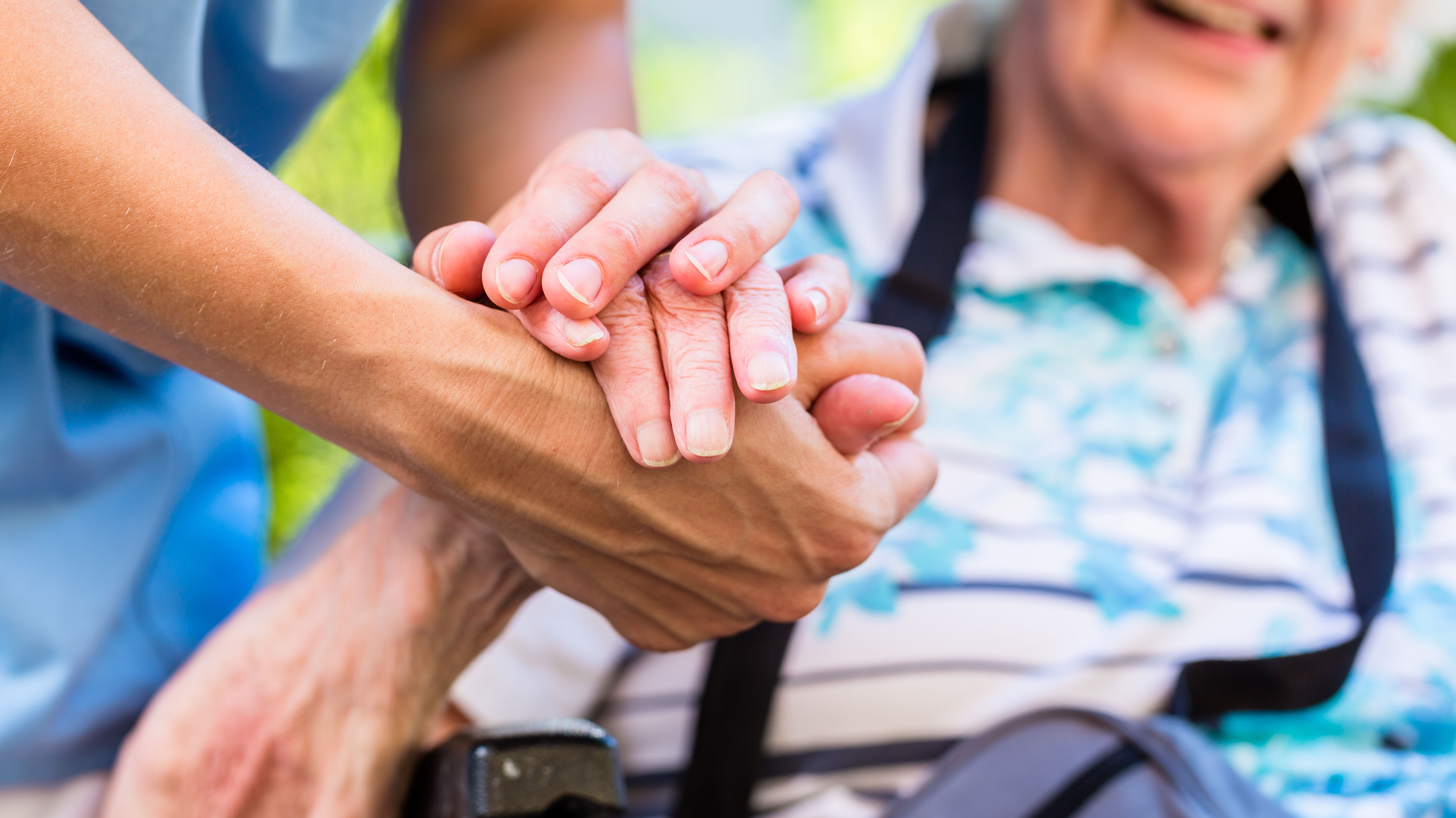 Die Hand eines Pflegers oder einer Pflegerin hält die Hand einer Seniorin im Rollstuhl.