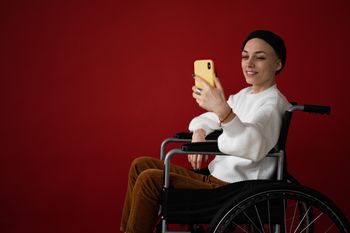 Eine junge Frau mit Wollmütze und lackierten Fingernägeln sitzt in einem Rollstuhl und schaut auf ihr Smartphone, dabei lächelt sie.