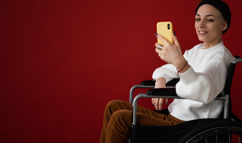 Eine junge Frau mit Wollmütze und lackierten Fingernägeln sitzt in einem Rollstuhl und schaut auf ihr Smartphone, dabei lächelt sie.
