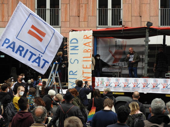 Ein Mann redet auf einer Bühne auf einer Demonstration davor weht eine weiße Fahne mit dem Logo "Parität"