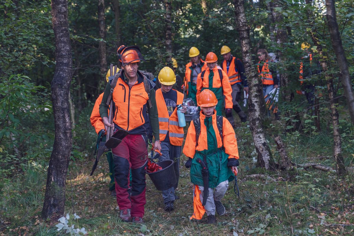 Copyright: Bergwald e. V. Das Bild zeigt eine erwachsene Person und mehrere Kinder in organener Schnittschutzkleidung im Wald.