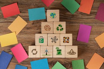 In der Mitte sind Holzwürfel auf denen Nachhaltigkeitssymbole wie Windkraft oder Recycling abgebildet sind. Außemrum liegen bunte Steine in Farben der SDGs. 
