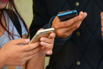 Zwei Personen, die beieinander stehen und jeweils auf ihrem Smartphone tippen. Der Fokus liegt auf den Händen und den Smartphones.