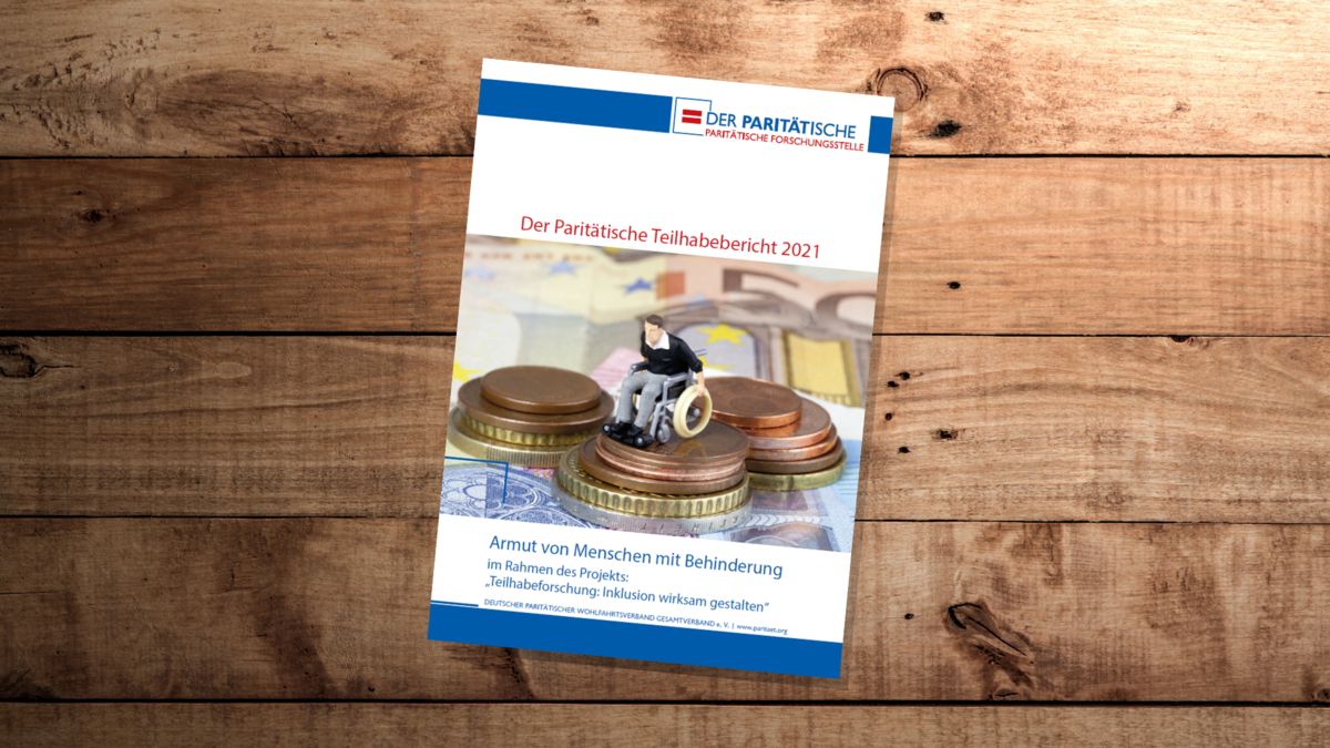 Auf einem Holztisch liegt eine Broschüre. Cover einer Broschüre. Auf dem Titelblitt sitzt eine kleine Figur im Rollstuhl auf einem Stapel Münzen. Überschrift der Broschüre: Der Paritätische Teilhabebericht 2021. Armut von Menschen mit Behinderung.