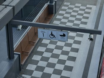 Zu sehen ist ein Bahnsteig von oben, der Boden besteht aus schwarz-weiß karierten Fliesen, an einem Metallbalken hängen Schilder, die die Nummer des Gleises zeigen sowie Piktogramme eines Rollstuhls und eines Kinderwagens. Auf dem Bahnsteig sind keine Personen zu sehen.