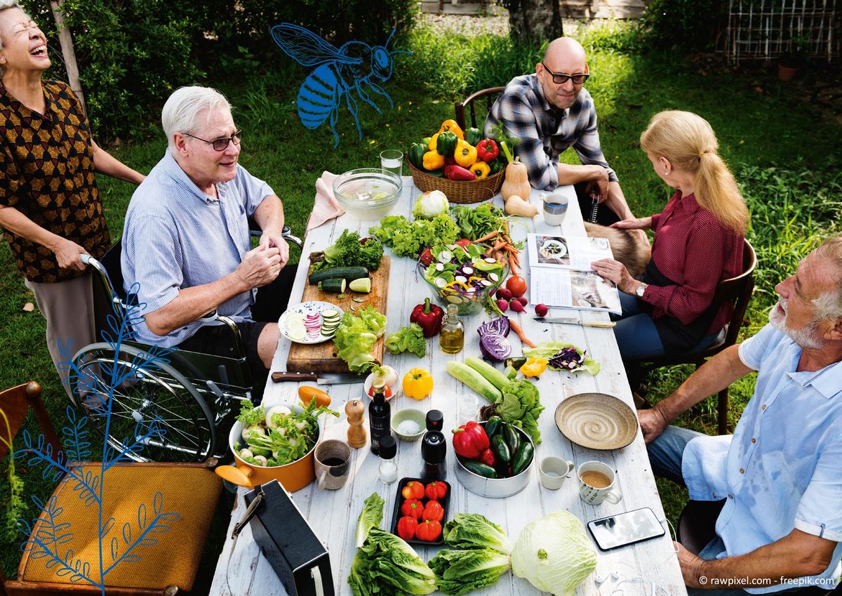Das Bild zeigt eine Gruppe älterer Menschen, zwei von ihnen mit Mobilitätsbeeinträchtigung, die im Freien an einem großen Tisch sitzen und frisches Gemüse zubereiten