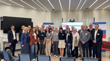 Ein Gruppenfoto aller Teilnehmerinnen und Teilnehmer der Tagung vor der Bildschimwand im Tagungsraum, darunter viele lächelnde Personen unterschiedlichen Alters in formeller Kleidung.