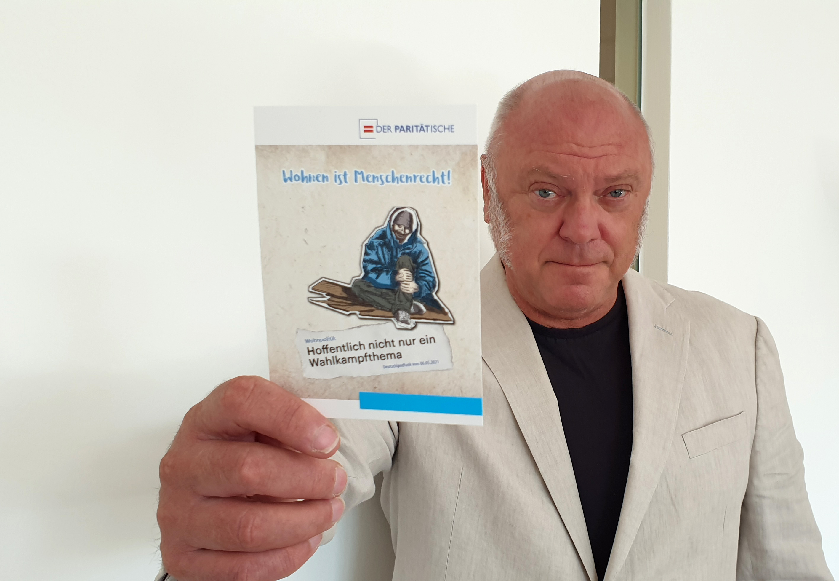 Ulrich Schneider, Hauptgeschäftsführer des Paritätischen Gesamtverbandes, hält eine Postkarte der Kampagne "Geh wählen, weil ALLE zählen" in die Kamera, die den Slogan "Wohnen ist Menschenrecht" zeigt.