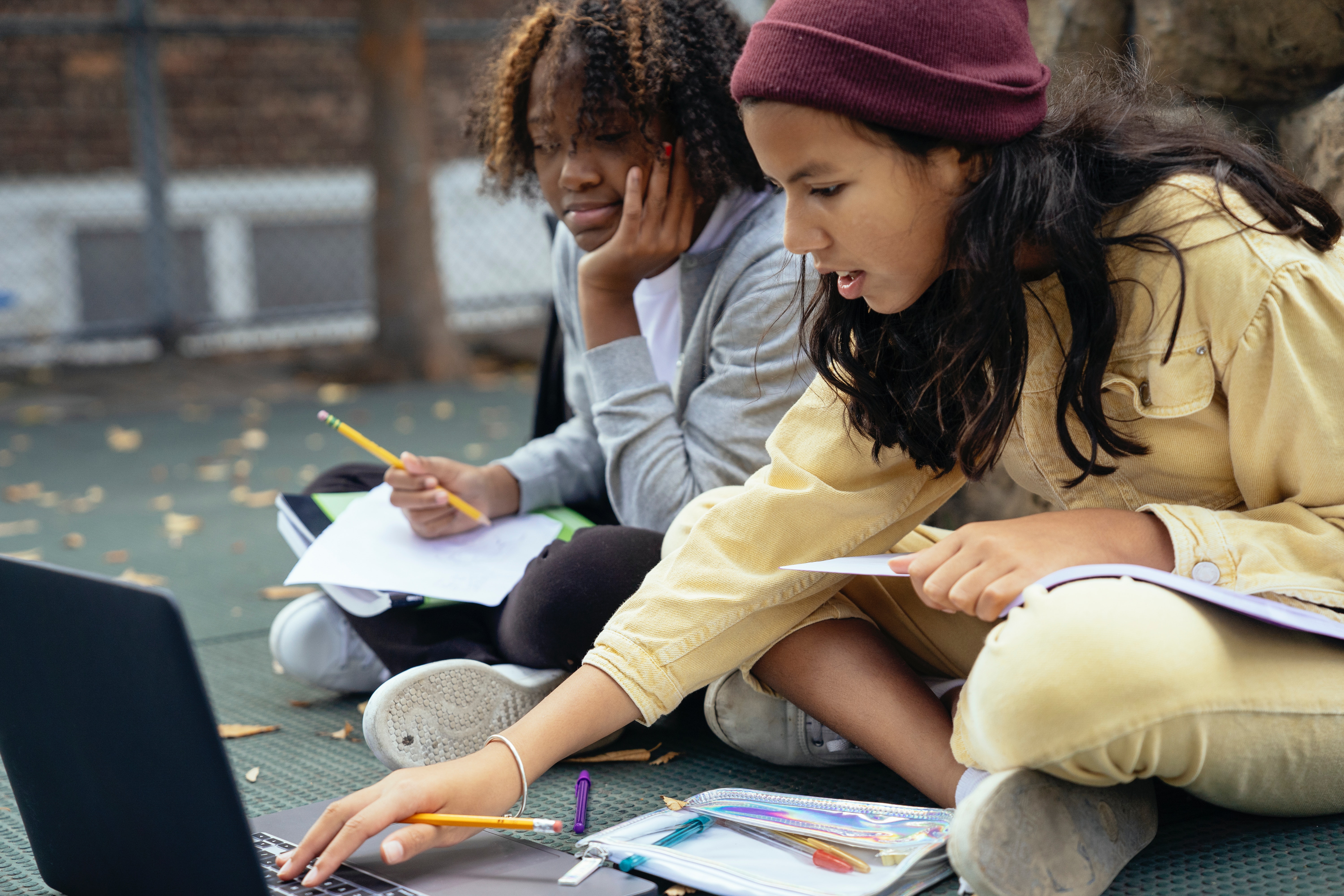 Zwei jugendliche Mädchen sitzen mit Stiften und Papier vor einem Laptop und erledigen Hausaufgaben. 