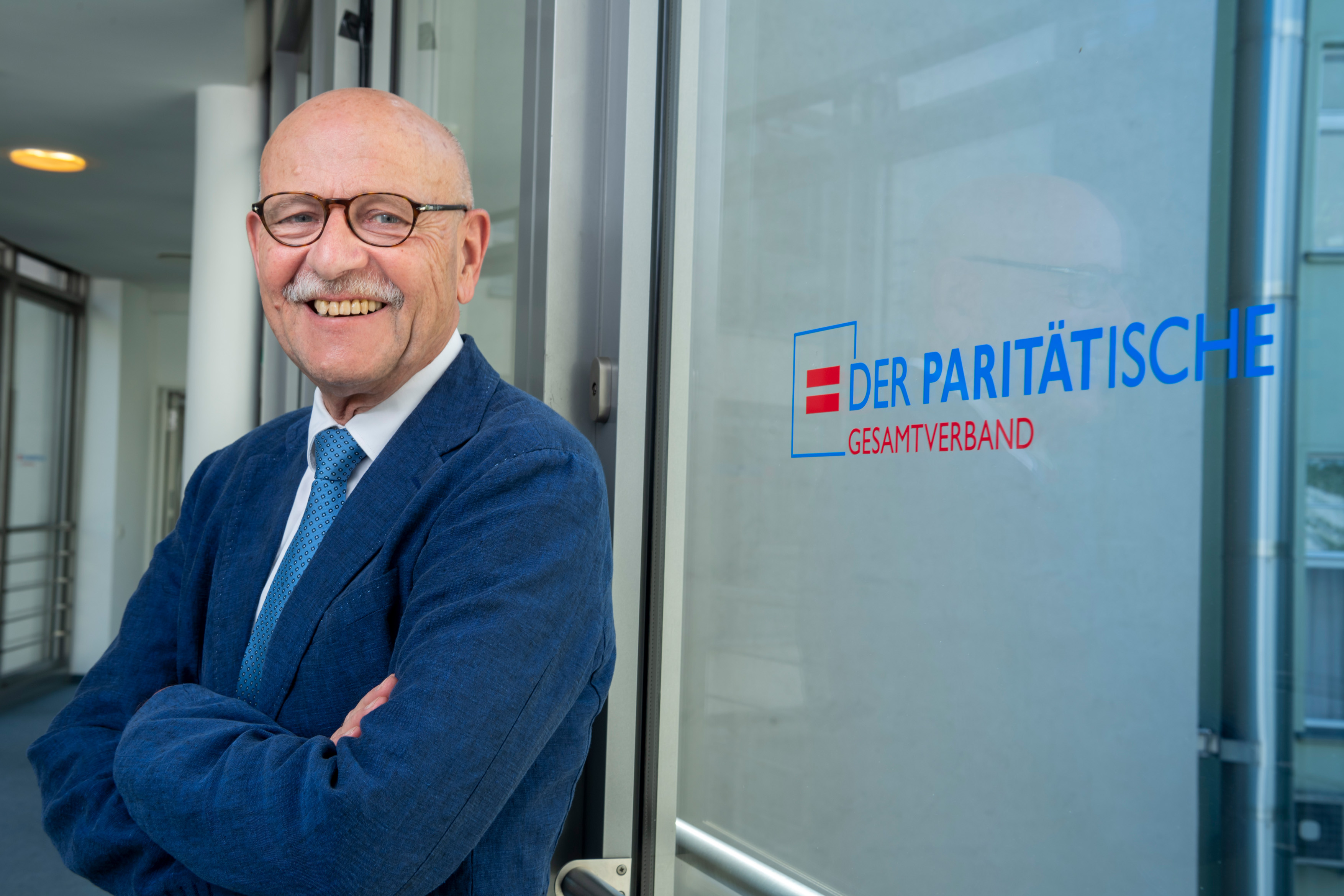 Ein Mann, mit Brille steht lächelnd vor einer Fensterscheibe, auf welcher das Logo des Paritätischen zu sehen ist. Es ist Prof. Dr. Rosenbrock, Vorsitzender des Paritätischen Gesamtverbandes.